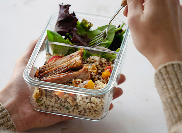 Eine Person hält eine durchsichtige Glasbox mit einem Mittagessen. Darin befinden sich Quinoa, bunte Gemüsewürfel und ein Stück gebratenes Fleisch. Zusätzlich liegt frischer grüner Salat obenauf. Eine Gabel ist in der anderen Hand der Person.