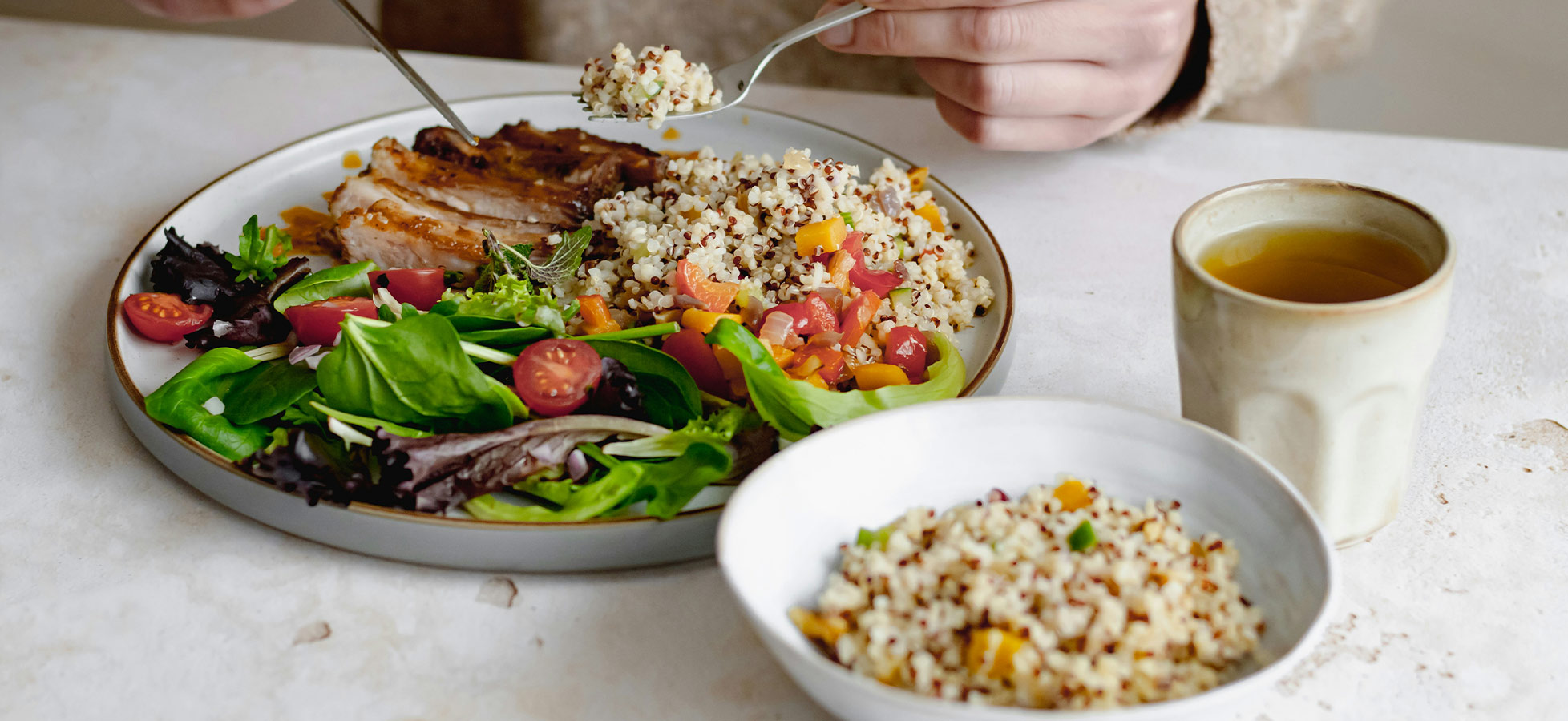 Eine Person isst von einem Teller mit gegrilltem Hähnchen, Quinoa-Salat und frischem Gemüse. Neben dem Teller steht eine kleine Schüssel mit mehr Quinoa und eine Tasse Tee. Der Fokus liegt auf der gesunden, bunten Mahlzeit.