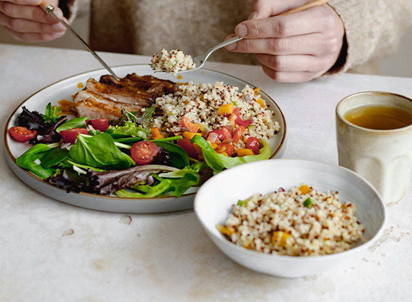Eine Person isst von einem Teller mit gegrilltem Hähnchen, Quinoa-Salat und frischem Gemüse. Neben dem Teller steht eine kleine Schüssel mit mehr Quinoa und eine Tasse Tee. Der Fokus liegt auf der gesunden, bunten Mahlzeit.