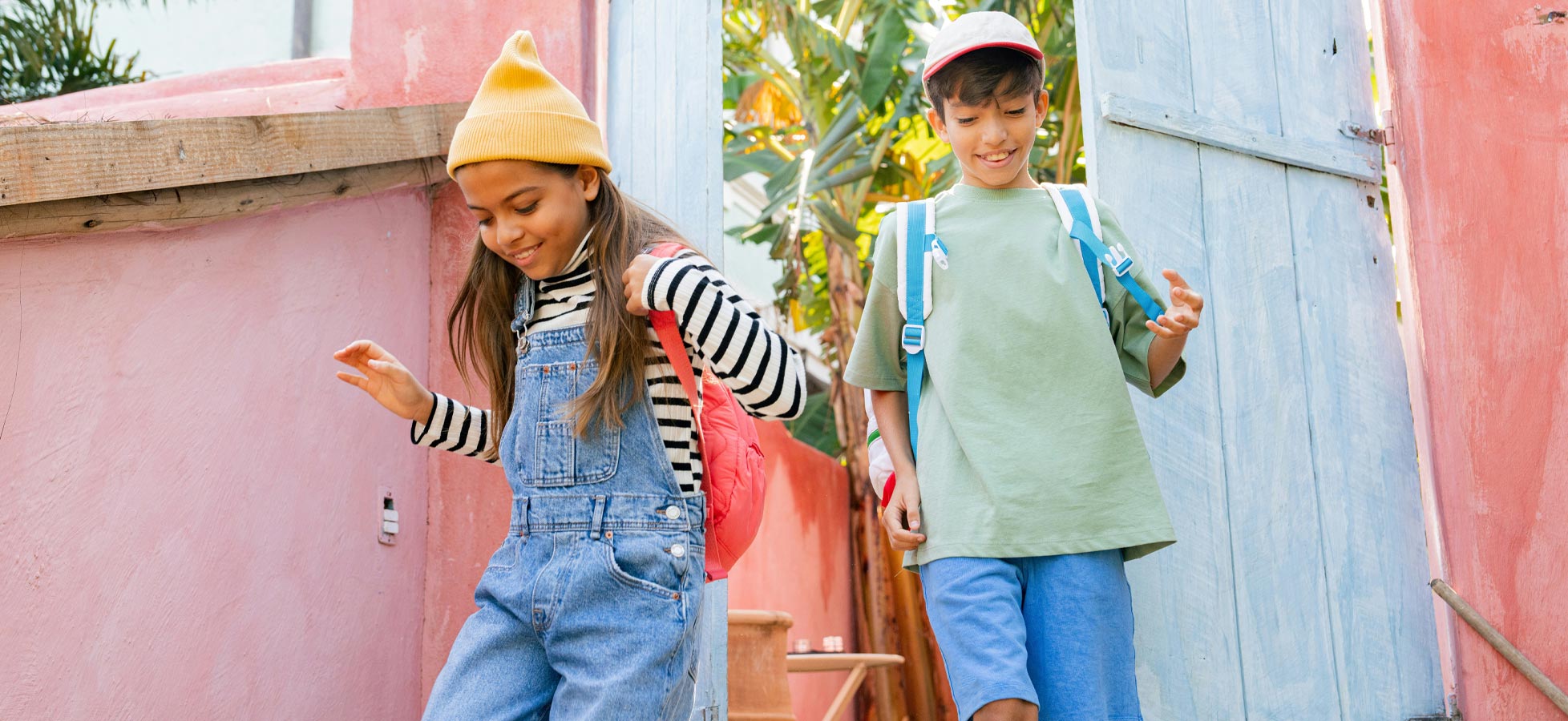 Zwei Kinder gehen fröhlich durch eine bunte, offene Tür. Das Mädchen trägt einen gestreiften Pullover und Latzhosen, während der Junge ein grünes T-Shirt und eine Mütze hat. Beide haben Rucksäcke auf den Schultern und lächeln, während sie miteinander interagieren.