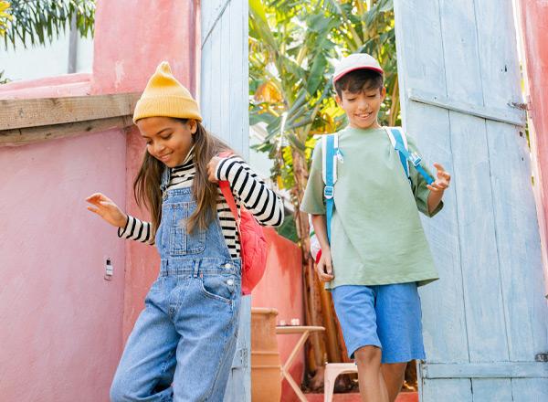 Zwei Kinder gehen durch ein blaues Tor. Ein Mädchen mit einem gelben Beanie und einem roten Rucksack trägt einen gestreiften Pullover und Jeans. Ein Junge mit einer weißen Kappe hat einen grünen T-Shirt und kurze Hosen an. Der Hintergrund zeigt eine lebendige, grüne Pflanzenlandschaft.