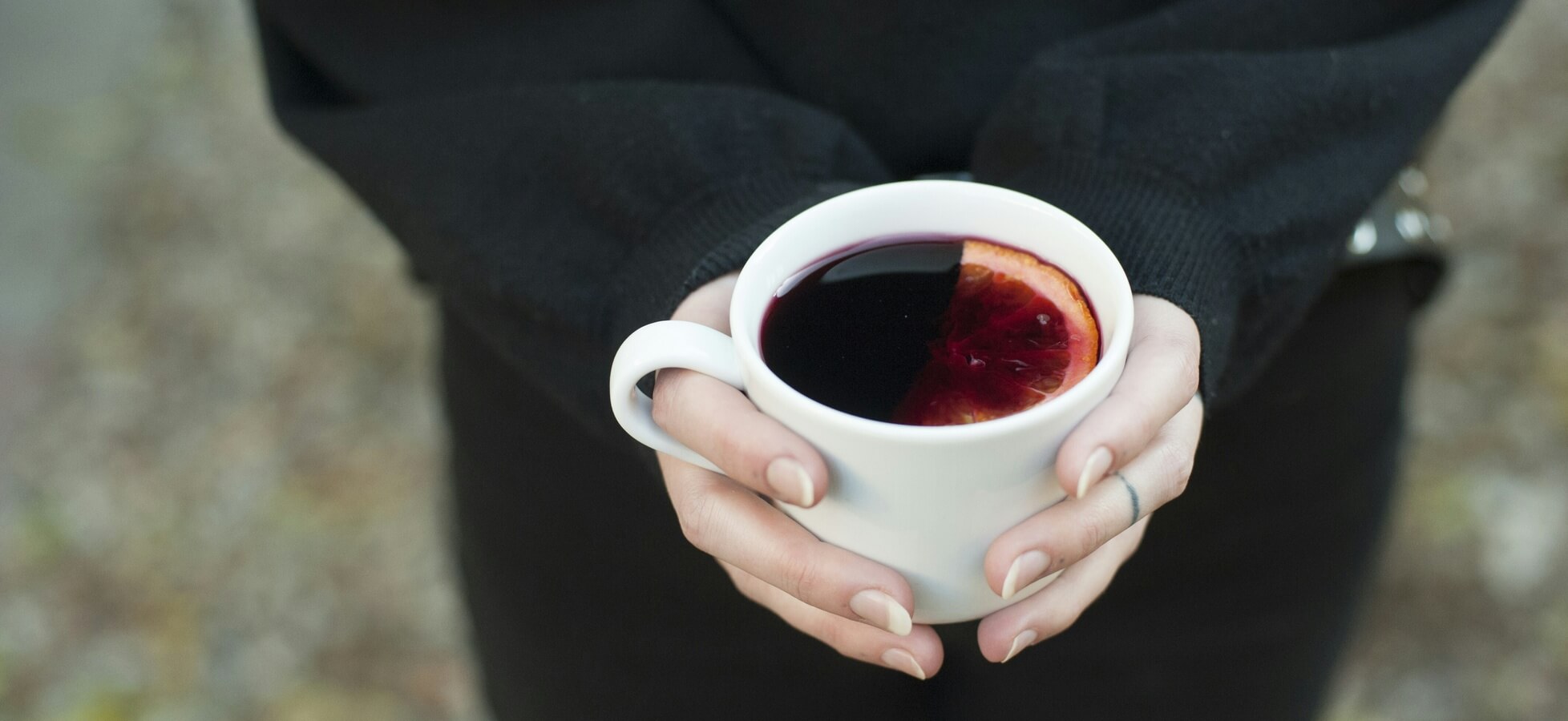 Eine Person hält eine weiße Tasse mit einem dunklen Getränk. An der Oberfläche des Getränks schwimmt eine halbe Blutorange, die dem Bild eine frische Note verleiht. Die Hände sind gut sichtbar und tragen einen schlichten, dunklen Pullover.