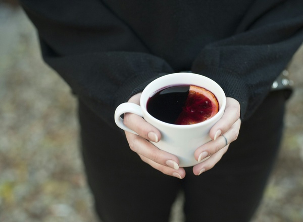 Eine Person hält eine weiße Tasse mit einem dunklen Getränk, das eine orangefarbene Scheibe Zitrusfrucht enthält. Die Person trägt einen schwarzen Pullover und die Hände sind sichtbar. Der Hintergrund ist unscharf und zeigt den Boden.