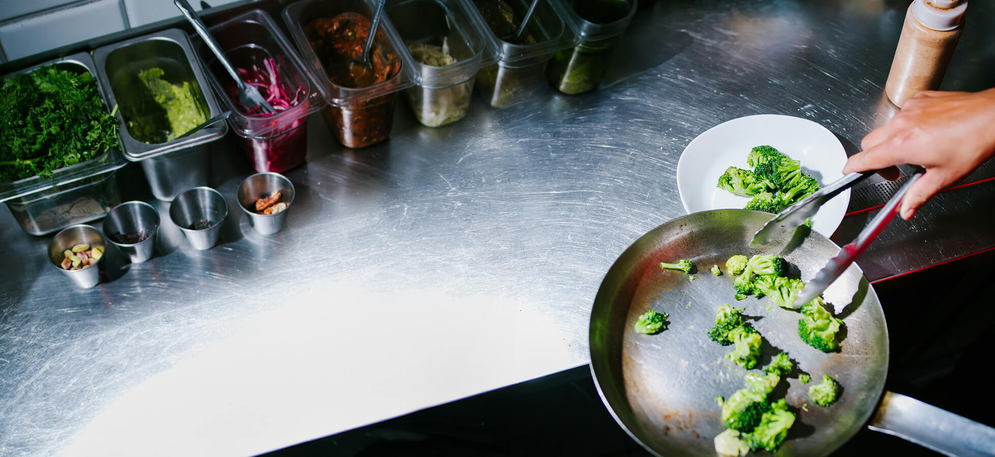 Eine Hand mit einer Zange hebt Brokkoli aus einer Pfanne. Im Hintergrund befinden sich verschiedene Zutaten in Behältern. Ein weißer Teller steht bereit, um das Gemüse aufzunehmen.
