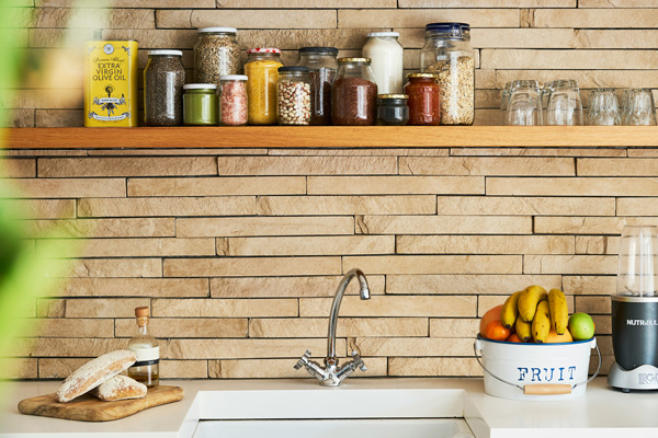 Eine moderne Küche mit einer Holzregal, das mehrere Gläser mit Lebensmitteln und Gewürzen enthält. Darunter ist eine Spüle mit einem Wasserhahn, neben der ein Obstkorb mit Bananen steht. Der Raum hat eine offene, freundliche Atmosphäre mit Steinwänden.