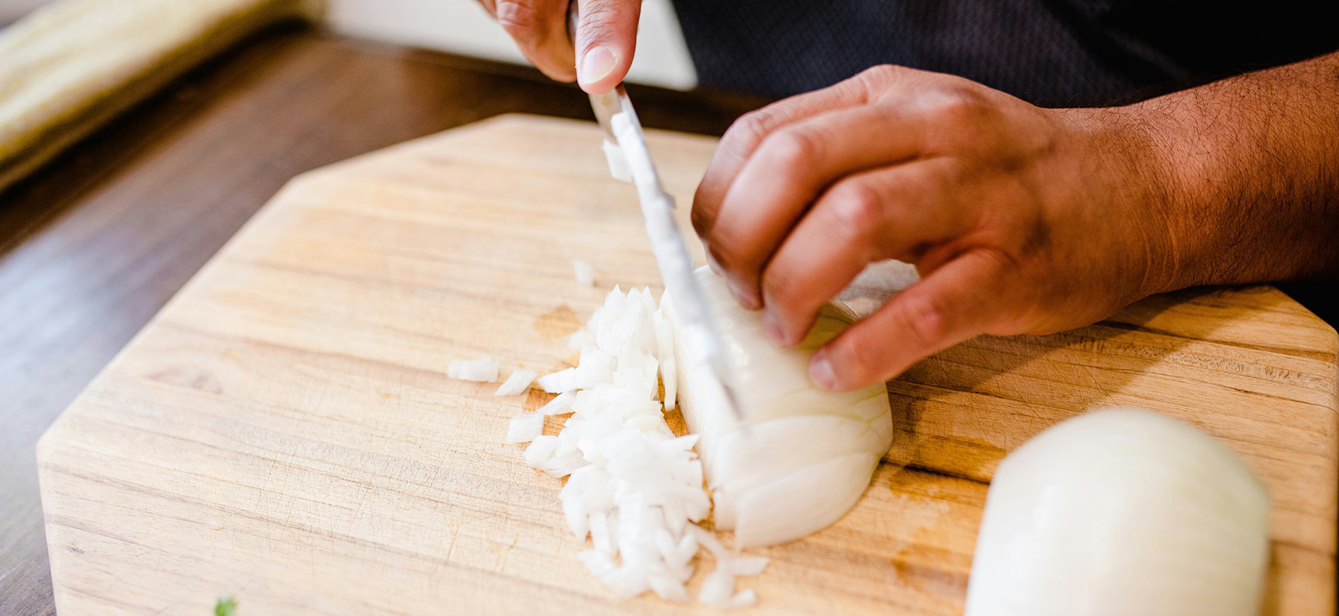 Eine Hand schneidet eine Zwiebel auf einem Holzbrett in kleine Stücke. Die Zwiebel ist halbiert und liegt auf der Schneidefläche. Der Fokus liegt auf dem Schnittprozess, während das Küchenumfeld unscharf im Hintergrund bleibt.
