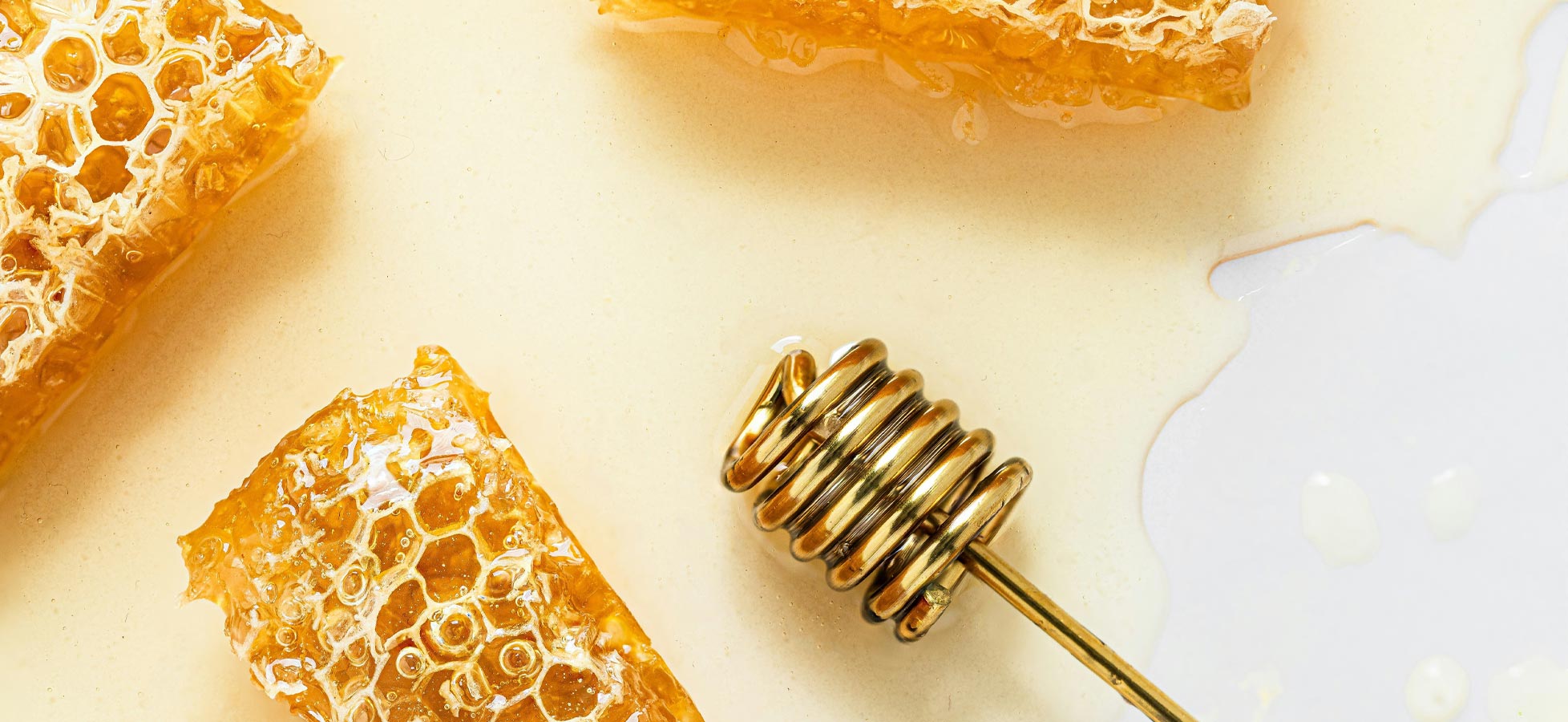 Drei Wabenstückchen aus Honig neben einem Honiglöffel, der golden glänzt. Der Honig fließt sanft und bildet kleine Tropfen auf einer hellen Unterlage. Die Waben haben eine durchscheinende, goldene Farbe, die die Natürlichkeit des Produkts betont.