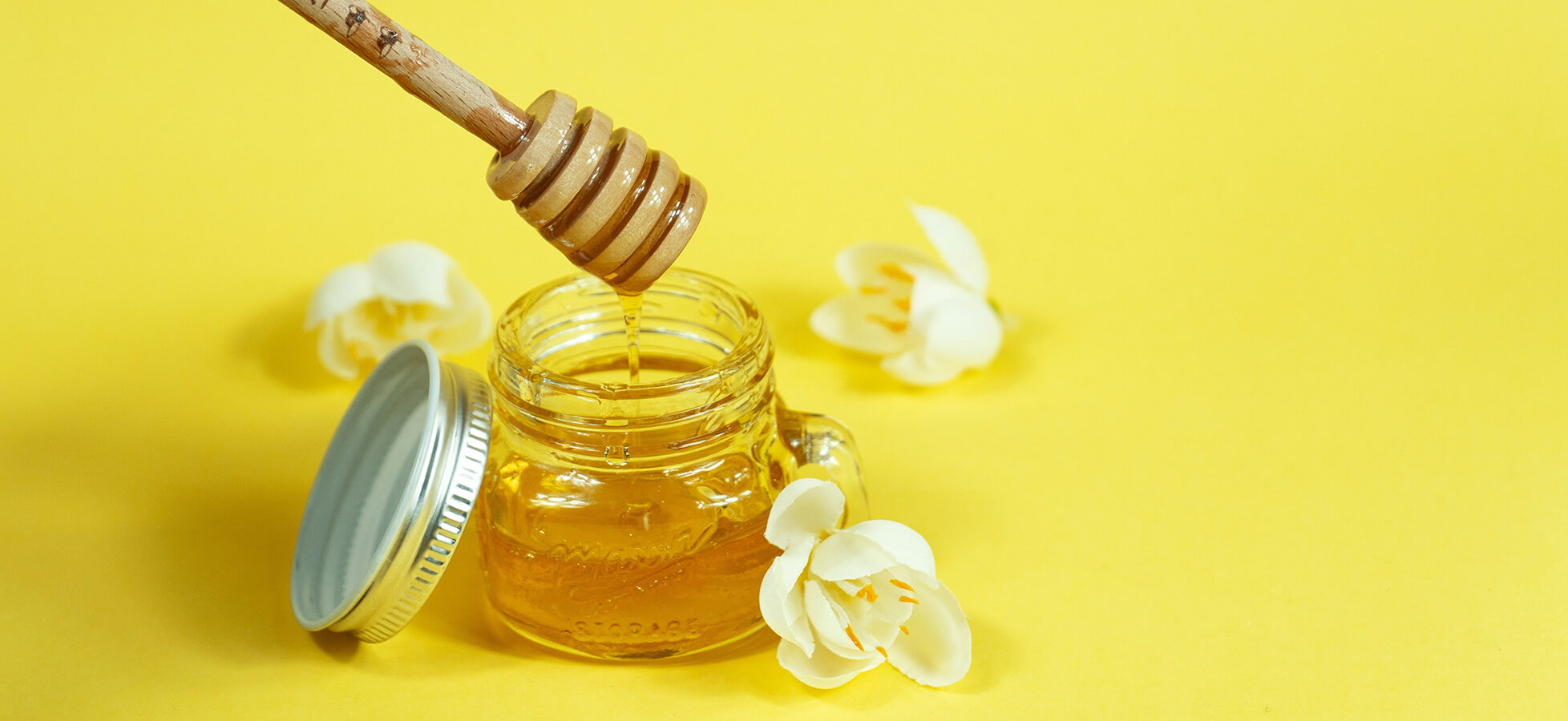 Ein Glas Honig steht auf einem gelben Hintergrund. Ein Honiglöffel mit einer runden Kante ist teilweise im Honig eingetaucht. Um das Glas liegen einige weiße Blumen.