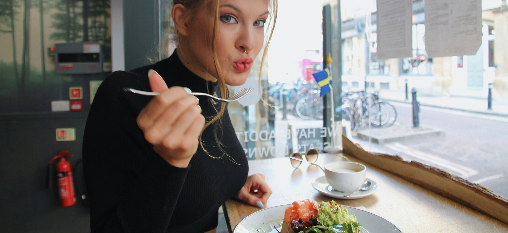 Eine junge Frau sitzt am Tisch und hält eine Gabel in der Hand. Sie schaut direkt in die Kamera und hat einen fordernden Gesichtsausdruck. Vor ihr steht ein Teller mit buntem Essen und eine Tasse Kaffee. Im Hintergrund sind Fahrräder und Fenster zu sehen.