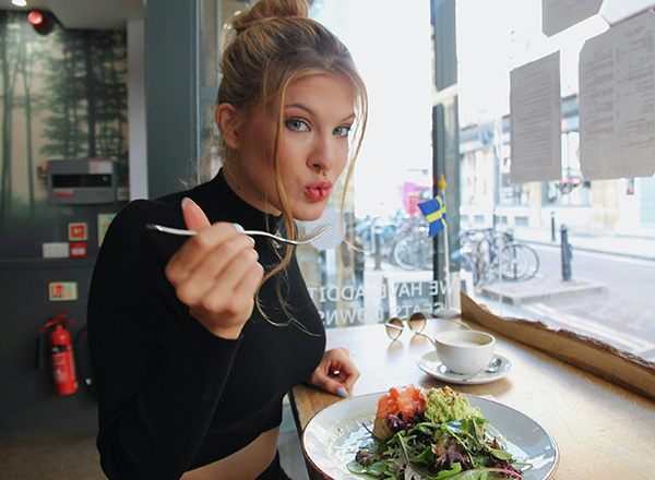 Eine junge Frau sitzt an einem Tisch und hält eine Gabel in der Hand. Sie lächelt und scheint einen Salat zu genießen, der auf dem Tisch vor ihr angerichtet ist. Neben dem Salat steht eine Tasse Kaffee. Hinter ihr ist ein Fenster mit Blick auf eine Straße.
