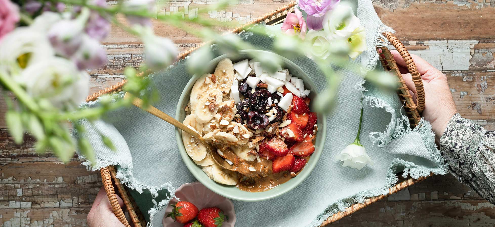 Eine handgehaltene Schale mit Müesli ist auf einem dekorativen Tablett platziert. Das Müesli ist mit Bananen, Erdbeeren, Kokosnuss und Nüssen garniert. Frische Blumen im Hintergrund ergänzen das ansprechende Arrangement.
