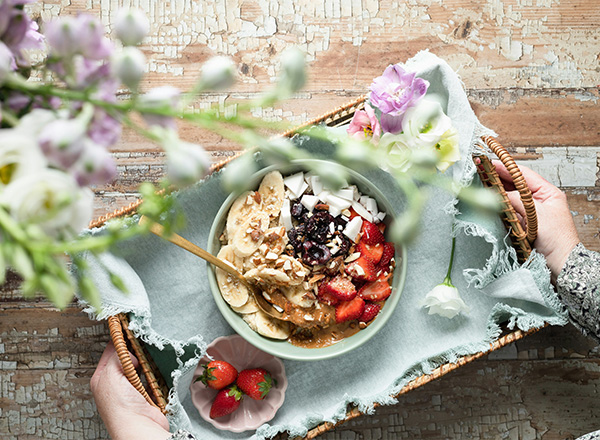 Eine Hand hält einen Korb mit einer Schüssel voller gesundem Frühstück: griechischer Joghurt, Bananen, Erdbeeren, Schokolade und Nüssen. Der Korb ist mit einer dezenten Stoffserviette ausgelegt und wird von blühenden Pflanzen umgeben.