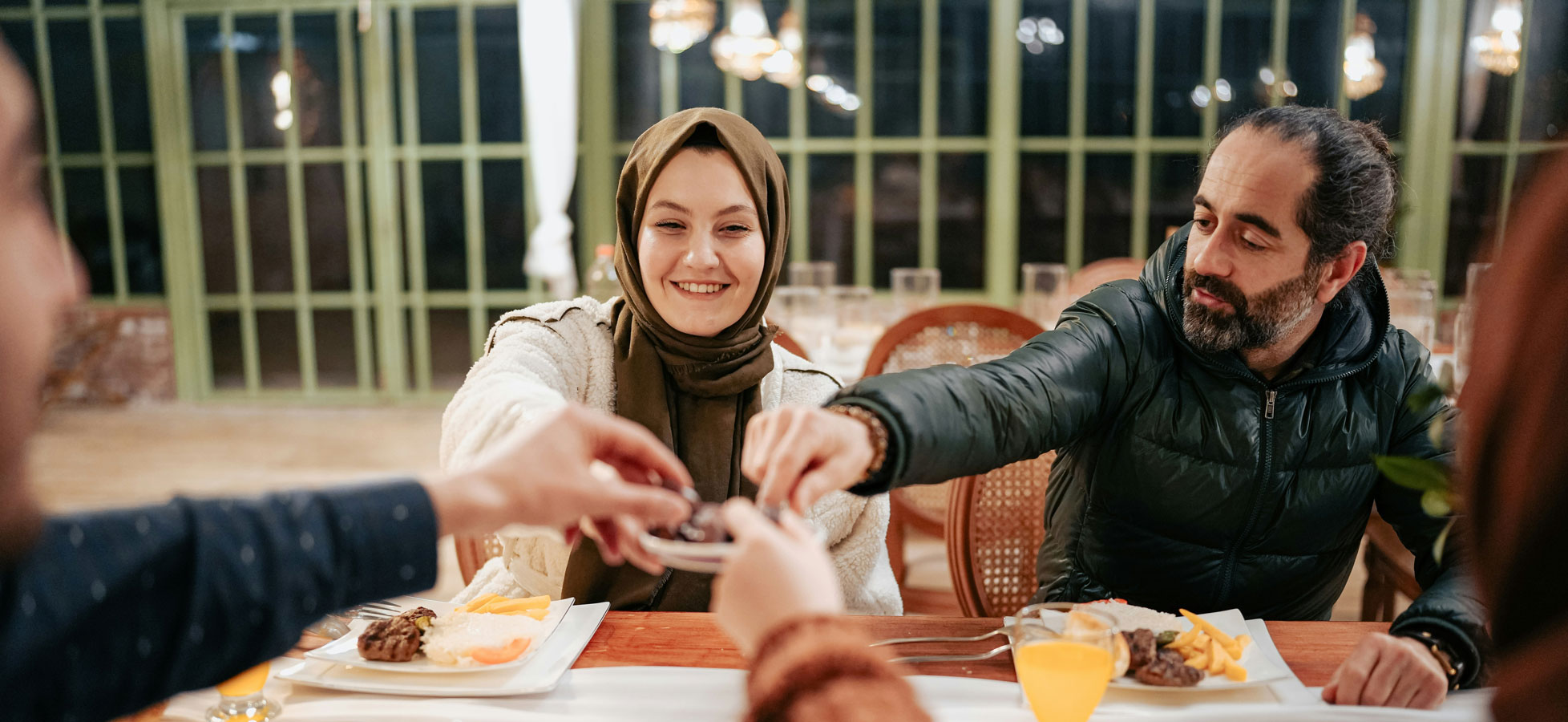 Eine Frau in einem Hijab sitzt auf einem Gebetsteppich in einem ruhigen Raum. Sie betet mit erhobenen Händen, während sanftes Licht durch dekorative Fenster auf den Boden fällt. Neben ihr liegen ein paar Bücher und Gebetsgegenstände.