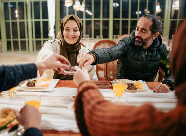 Eine Frau in traditioneller islamischer Kleidung sitzt auf dem Boden einer Moschee und betet. Vor ihr liegt ein Gebetsteppich mit dem Koran und anderen religiösen Gegenständen. Im Hintergrund sind dekorative Fenster mit geometrischen Mustern zu sehen, die Licht auf den Raum werfen.