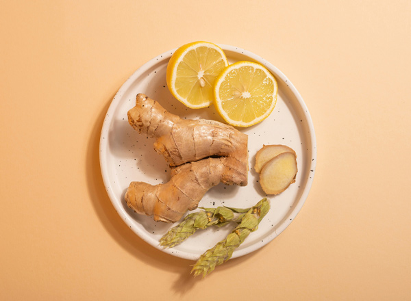 Auf einem weißen Teller liegen frische Ingwerstücke, eine geschnittene Limone und einige grüne Spargelspitzen. Der Hintergrund ist in sanftem Beige gehalten, was den Fokus auf die Naturprodukte lenkt.