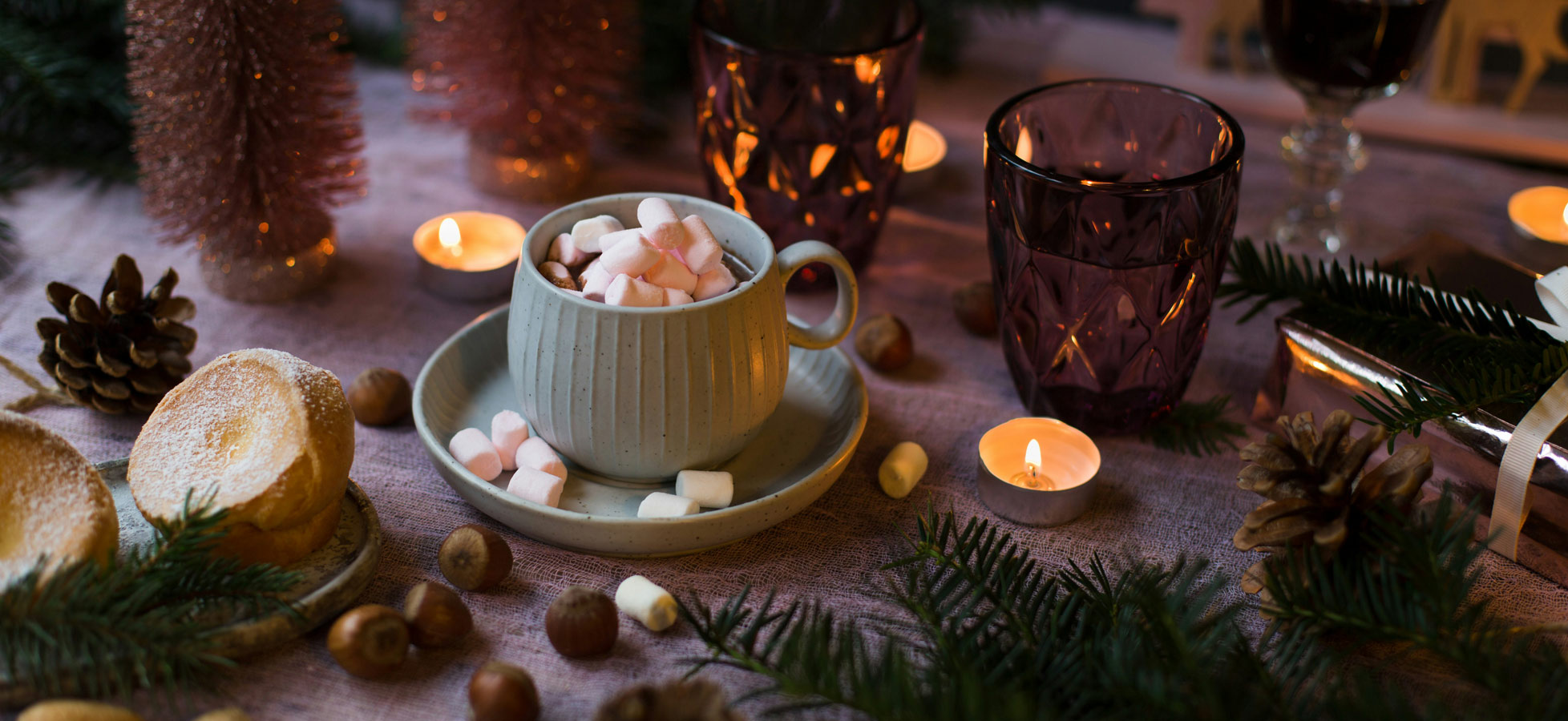 Eine gemütliche Tischszene mit einer Tasse, gefüllt mit bunten Mini-Marshmallows, auf einem Unterteller. Rundherum liegen Kekse, Nüsse und Tannenzweige. Es sind Kerzenleuchten und dekorative Weihnachtsbäumchen im Hintergrund, die eine festliche Atmosphäre schaffen.