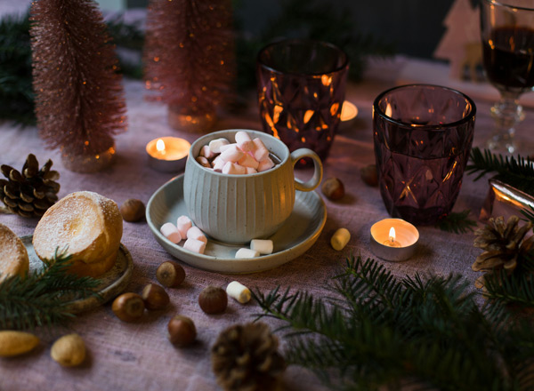 Eine gemütliche Tischszene mit einer Tasse, gefüllt mit bunten Mini-Marshmallows, auf einem Unterteller. Rundherum liegen Kekse, Nüsse und Tannenzweige. Es sind Kerzenleuchten und dekorative Weihnachtsbäumchen im Hintergrund, die eine festliche Atmosphäre schaffen.