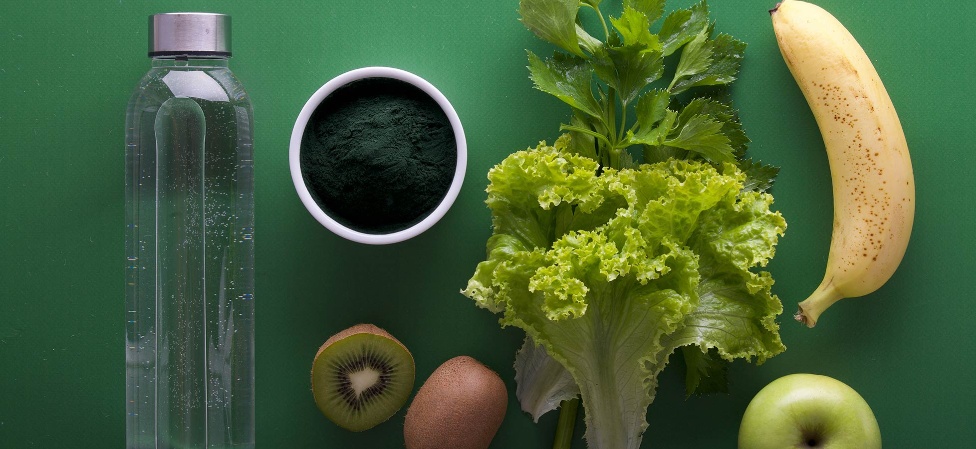 Ein Erfrischungsbild mit einer Wasserflasche, einer Schüssel mit Spirulina-Pulver, frischem Salat, Sellerie, einer Banane, einem Kiwi, einer Kiwi und einem grünen Apfel auf einem grünen Hintergrund.