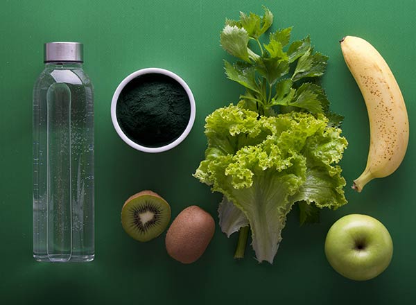 Das Bild zeigt eine Anordnung von Lebensmitteln auf einem grünen Hintergrund. Zu sehen sind ein Glas Wasser, eine Schüssel mit spirulina Pulver, frischer Blatt-Salat, eine Banane, ein Kiwi, ein brauner Kiwifruit und ein Apfel.