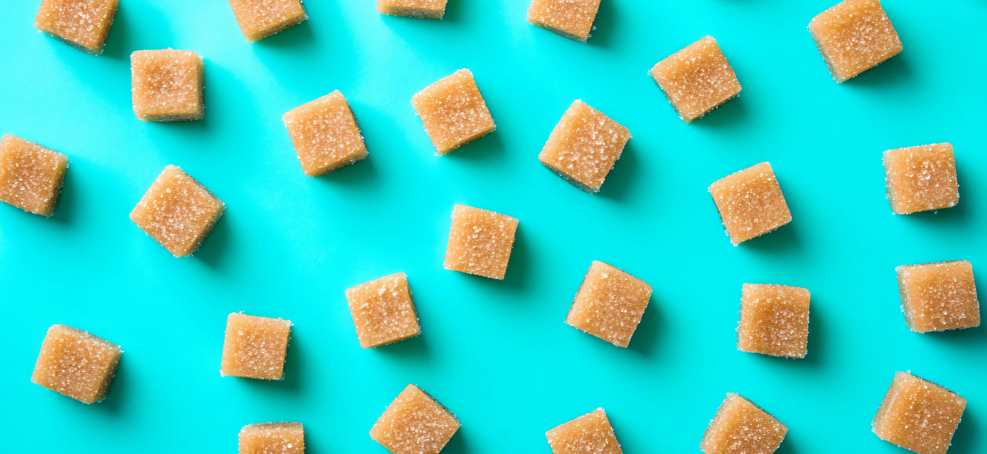 Quadratische, karamellfarbene Würfelzuckerwürfel liegen gleichmäßig verteilt auf einem aquablauen Hintergrund. Die Würfel sind leicht glitzernd und scheinen aus Zucker gefertigt zu sein.