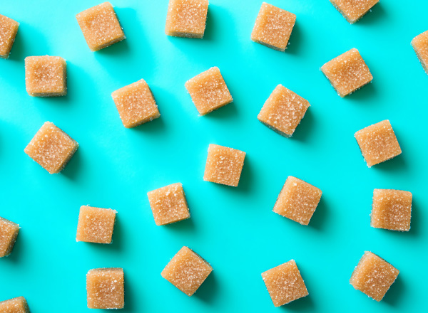 Auf einem hellblauen Hintergrund sind gleichmäßig verteilte, goldbraune Würfel aus Zucker zu sehen. Sie sind klein und haben eine glatte Oberfläche, mit einer leichten Zuckerschicht, die sie funkeln lässt. Das Bild vermittelt eine süße und einladende Atmosphäre.