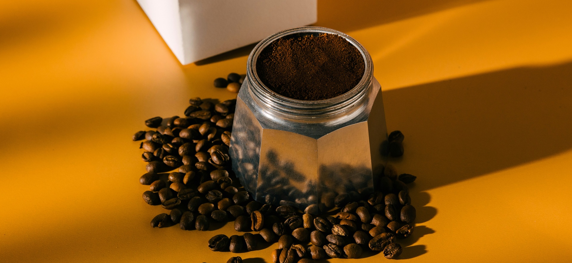 Ein Kaffeebereiter aus Metall steht auf einem orangefarbenen Tisch, umgeben von Kaffeebohnen. Der Deckel des Behälters ist teilweise geöffnet und zeigt frisch gemahlenen Kaffee. Es gibt eine dynamische Schattenbildung auf der Oberfläche.