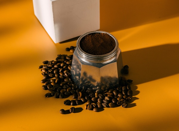 Eine Kaffeekanne aus Edelstahl steht auf einem gelben Tisch. Um sie herum liegen verstreut Kaffeebohnen, während der Deckel der Kanne halb geöffnet ist und gemahlener Kaffee sichtbar ist. Das Licht erzeugt sanfte Schatten.