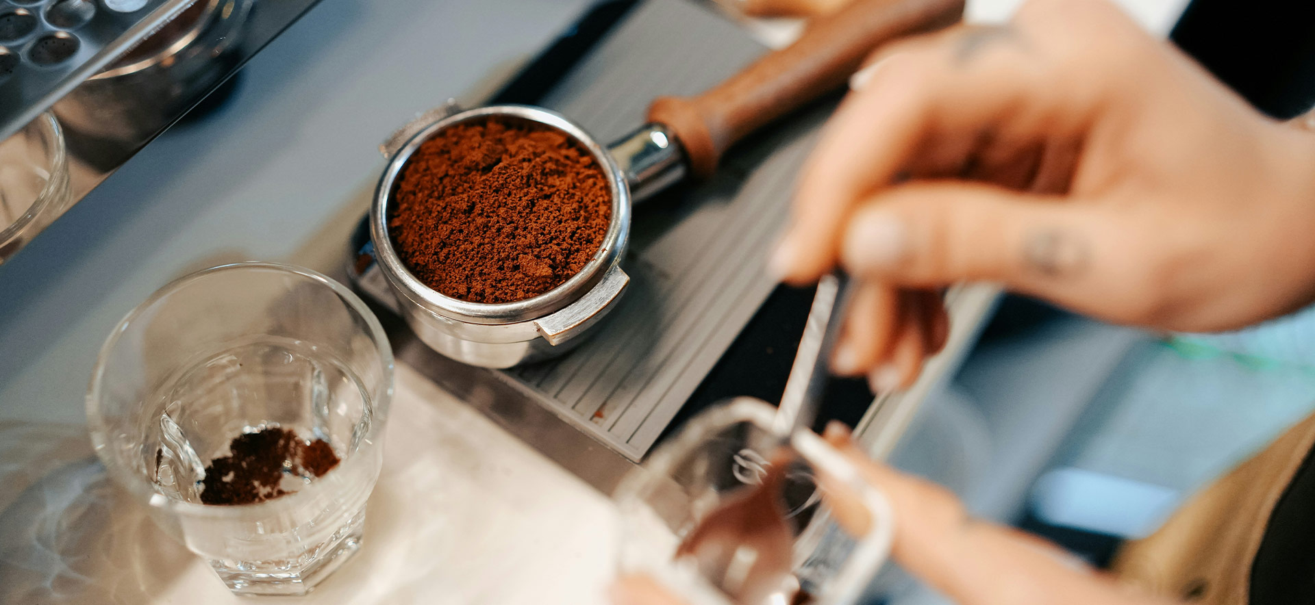 Eine Hand hält einen Messlöffel über ein Glas, während die andere Hand einen Siebträger mit gemahlenem Kaffee zeigt. Im Hintergrund ist ein Kaffeegerät und ein Glas Wasser zu sehen.