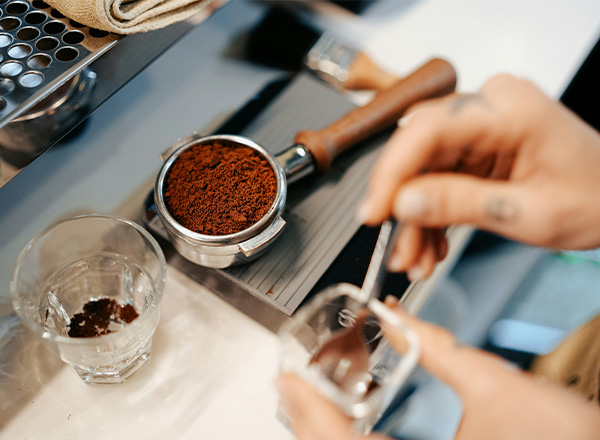 Eine Hand hält einen Messlöffel über ein Glas, während die andere Hand einen Kaffeepfilterträger mit gemahlenem Kaffee zeigt. Im Hintergrund ist eine Maschine für die Kaffeezubereitung sichtbar.