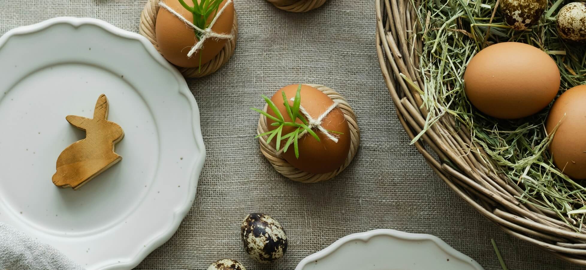 Eine rustikale Tischdeko zeigt verschiedene Eier, einige mit grünen Zweigen dekoriert, und einen kleinen Holzhasen. Ein geflochtener Korb enthält weitere Eier und befindet sich auf einer textilen Unterlage. Die Szene vermittelt eine festliche Osteratmosphäre.
