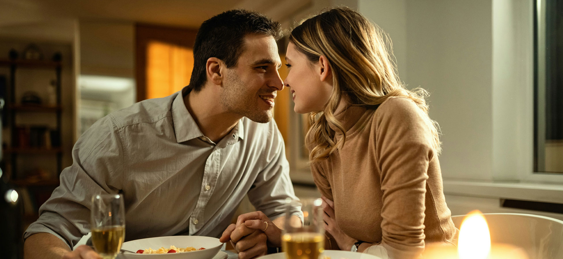 Ein Paar sitzt an einem Tisch, konzentriert aufeinander. Sie lächeln sich an und scheinen sich sehr nahe zu sein. Auf dem Tisch stehen Teller mit Essen und Gläser mit Getränken. Eine gemütliche, romantische Atmosphäre ist spürbar.
