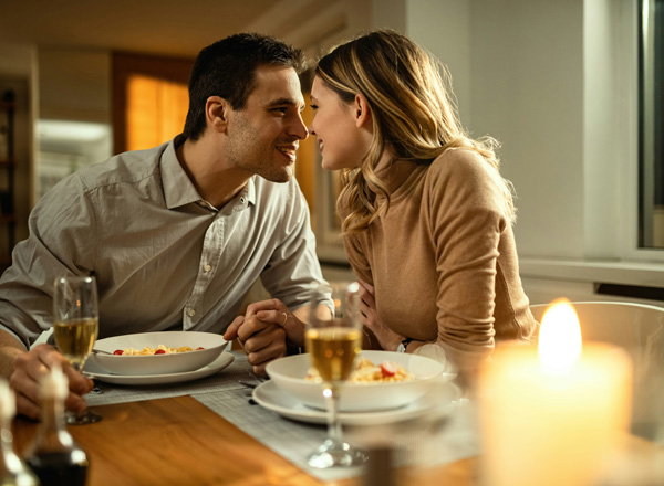 Ein Paar sitzt bei einem romantischen Abendessen am Tisch, blickt sich liebevoll in die Augen und lächelt sich an. Vor ihnen stehen Teller mit Pasta und Gläser mit Sekt. Ein Kerzenlicht sorgt für eine gemütliche Atmosphäre.