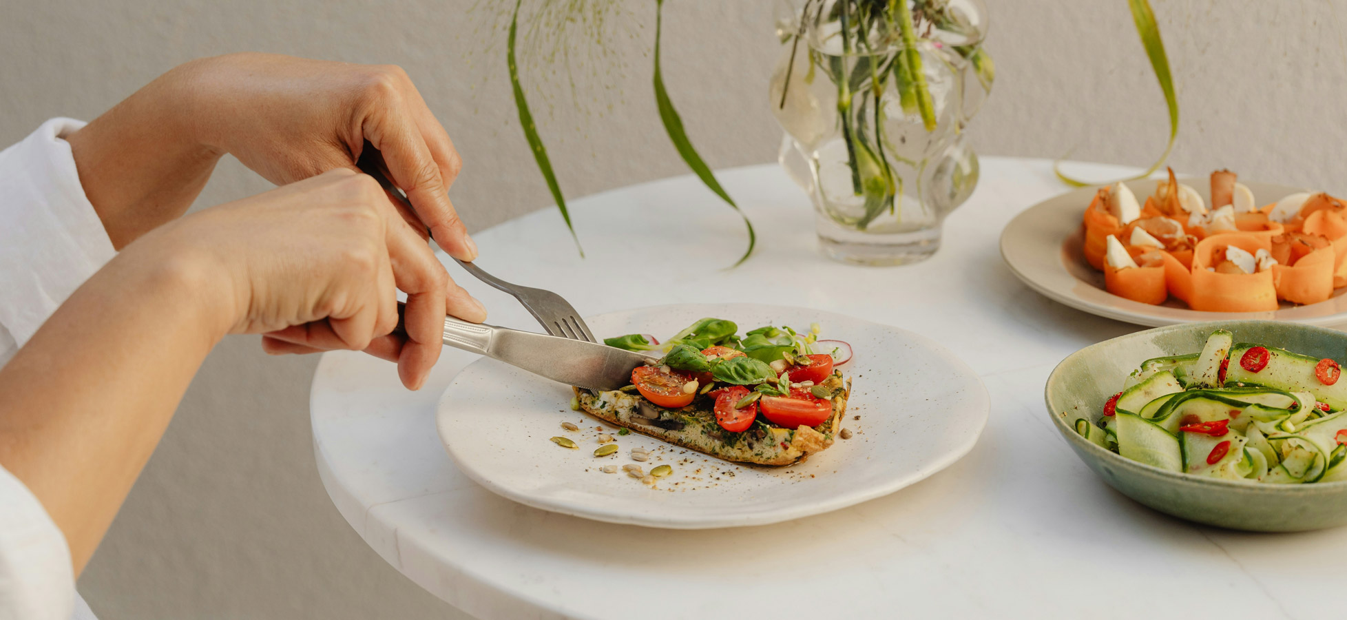 Eine Person schneidet mit Messer und Gabel ein Stück belegtes Brot mit Tomaten und Kräutern auf einem weißen Teller. Im Hintergrund sind weitere Gerichte, darunter gemischtes Gemüse, auf einem Tisch sichtbar.