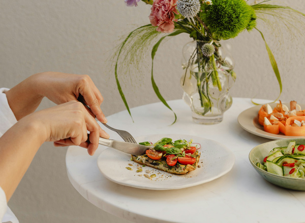 Eine Hand schneidet ein Stück gegrillten Fisch mit Tomaten und frischem Gemüse auf einem weißen Teller. Im Hintergrund sind verschiedene Salate und eine Blumenvase sichtbar. Der Tisch ist weiß und vermittelt eine helle, einladende Atmosphäre.