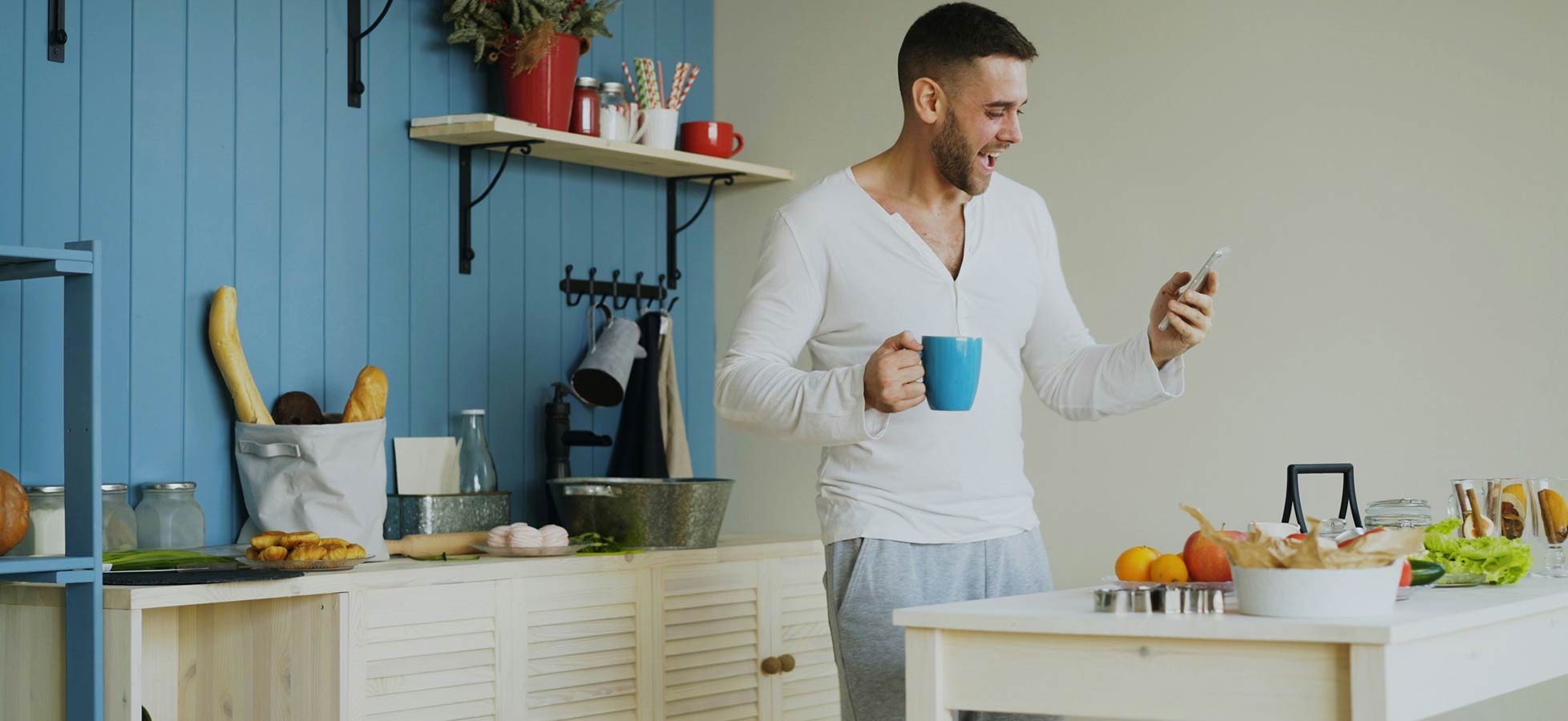 Ein Mann steht in einer hellen Küche, hält eine Kaffeetasse in einer Hand und schaut auf sein Smartphone. Auf der Kücheninsel sind frisches Gemüse und Obst zu sehen. Der Mann scheint erfreut oder interessiert zu sein.