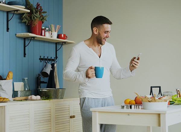 Ein Mann steht in einer modernen Küche, hält eine Tasse in der einen Hand und ein Smartphone in der anderen. Er lächelt, während er auf das Smartphone schaut. Auf dem Tisch sind frische Lebensmittel wie Obst und Gemüse sowie eine Wasserflasche zu sehen.