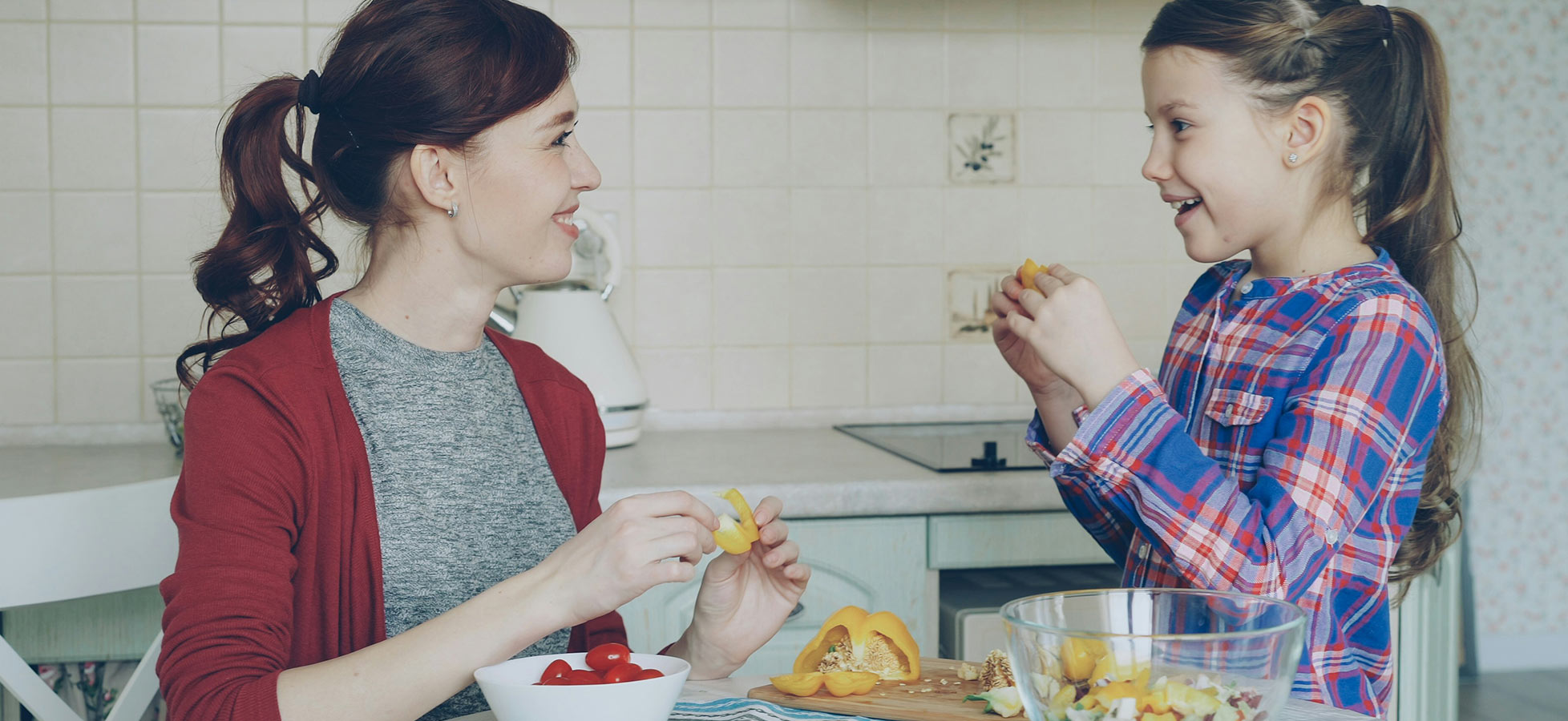 Eine Frau und ein Mädchen sitzen an einem Tisch in einer Küche. Sie lächeln sich an und teilen frisches Obst. Vor ihnen steht eine Schüssel mit Obst und eine Holzplatte mit weiteren Zutaten. Die Atmosphäre ist freundlich und gemeinschaftlich.