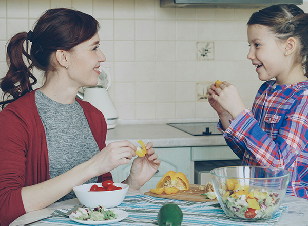 Eine Mutter und ihre Tochter sitzen an einem Tisch in der Küche und teilen Obst. Beide lächeln und genießen die Zeit miteinander, während sie gesunde Snacks zubereiten. Im Hintergrund sind frische Zutaten und eine Schüssel mit Salat zu sehen.