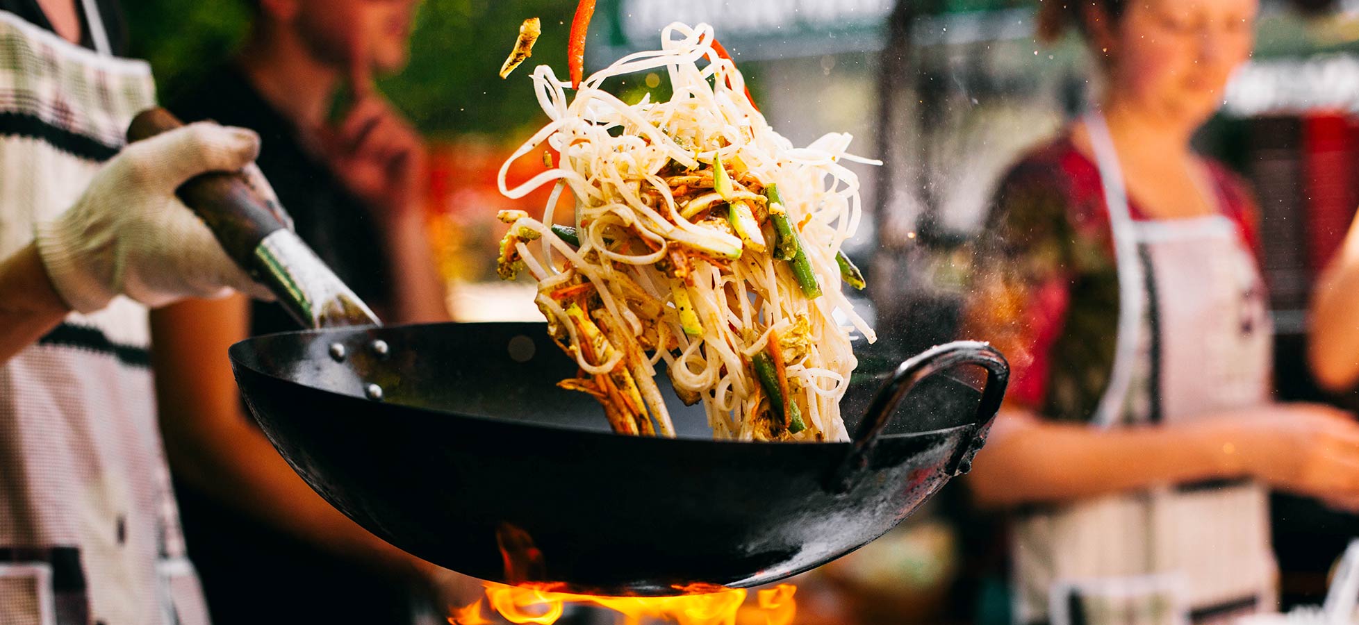 Eine Hand hält einen Wok, aus dem dampfende Nudeln mit Gemüse in die Luft schwingen. Im Hintergrund sind verschwommene Personen zu sehen, die ebenfalls in der Kochumgebung stehen. Flammen sind unter dem Wok sichtbar, was die Zubereitung von Speisen hervorhebt.