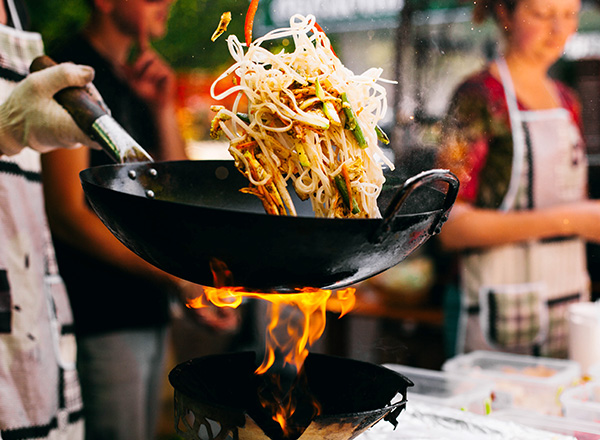 Ein Koch wirft gebratene Nudeln und Gemüse in eine wokähnliche Pfanne, während Flammen aufsteigen. Im Hintergrund sind weitere Personen sichtbar, die am Kochvorgang interessiert sind. Es herrscht eine lebhafte, gastronomische Atmosphäre.