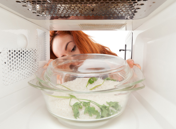 Eine Person mit roten Haaren schaut in die Mikrowelle, um eine Glasschüssel mit Reis und Kräutern zu überprüfen. Die Schüssel ist im Vordergrund sichtbar, während die Person sich näher nähert, um den Inhalt zu sehen.