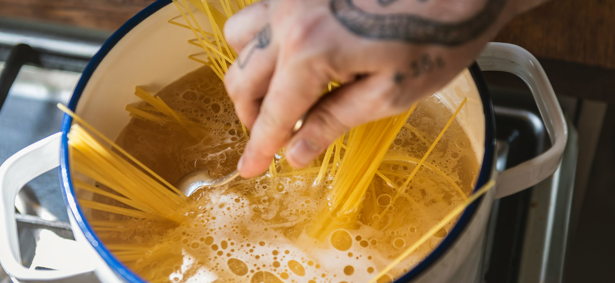 Ein Koch rührt Spaghetti in einem großen Topf mit kochendem Wasser. Blasen steigen auf und zeigen, dass das Wasser dampft. Die Hand des Kochs ist tätowiert und hält eine Gabel, während die langen Nudeln teilweise im Wasser stehen.