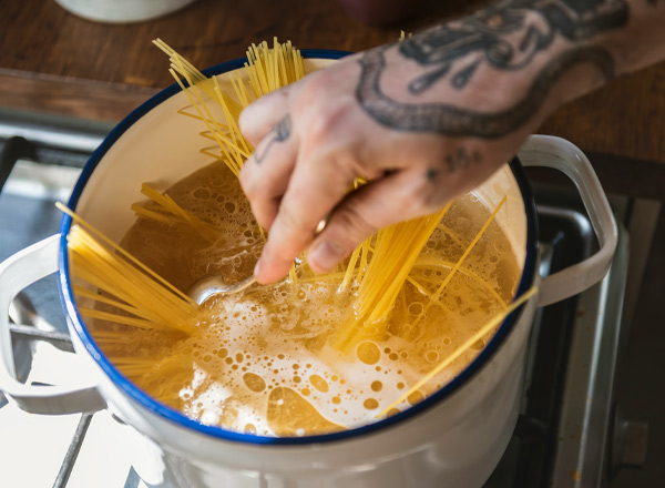 Ein Koch rührt Spaghetti in einem großen Topf mit kochendem Wasser. Blasen steigen auf und zeigen, dass das Wasser dampft. Die Hand des Kochs ist tätowiert und hält eine Gabel, während die langen Nudeln teilweise im Wasser stehen.