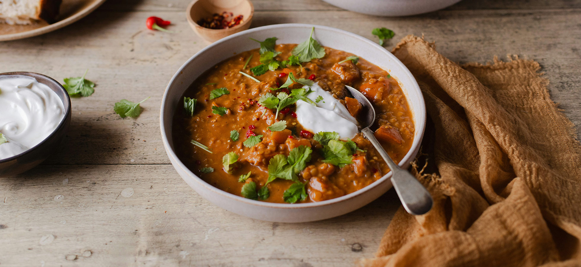 Zwei Schalen mit herzhaftem Eintopf sind auf einem Holztisch angerichtet. Der Eintopf ist mit frischem Koriander und einem Klecks Joghurt garniert, während rote Chilischoten und Gewürze daneben liegen. Im Hintergrund sind einige Stücke Brot sichtbar.