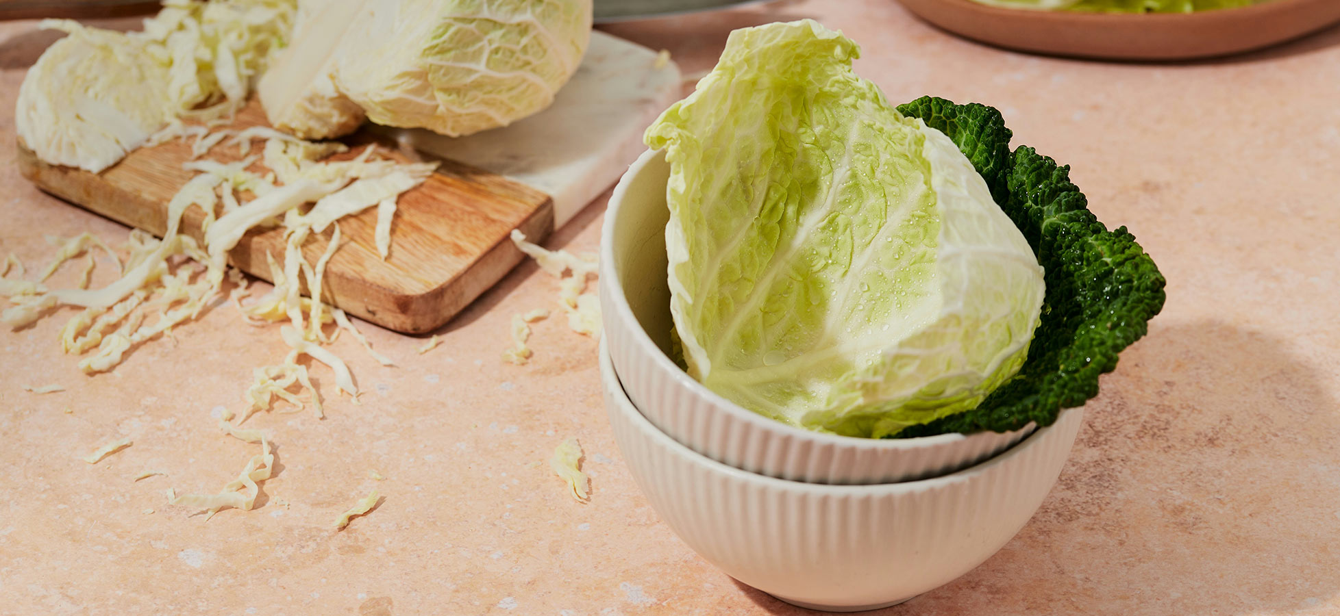 Eine weiße Schüssel enthält frische, grüne und weißliche Blätter von Kohl. Im Hintergrund liegt ein Holzbrett mit geschnittenem Kohl und verstreuten Kohlstreifen auf einer rosa Oberfläche.