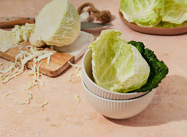 Eine Schüssel mit verschiedenen Blättern von Weißkohl und Wirsing steht auf einem Tisch. Neben der Schüssel liegt ein halbierter Kopf Weißkohl auf einem Holzbrett, umgeben von feinen Kohlstreifen. Der Hintergrund zeigt weiteren Kohl.