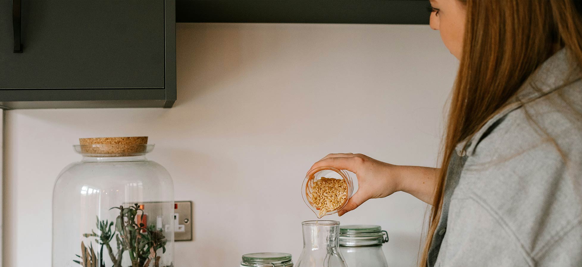 Eine Person mit langen Haaren füllt mit einem kleinen Glas Nüsse in ein Glasbehältnis auf einer Küchentheke. Im Hintergrund steht ein großes Glas mit Pflanzen. Die Küchenelemente sind modern und schlicht gestaltet.