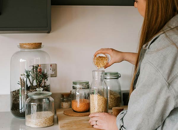 Eine Person füllt einen Glasbehälter mit einer Körnermischung in eine größere Glasflasche. Im Hintergrund stehen verschiedene Gläser mit unterschiedlichen Lebensmitteln auf einer Holzunterlage. Eine Pflanzenarrangement ist in einem großen Glasbehälter sichtbar.