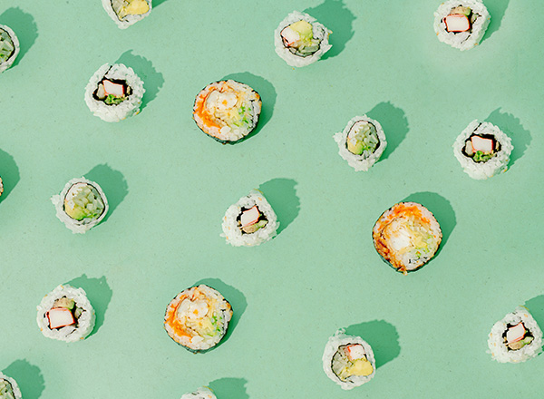 Auf einem mintgrünen Hintergrund sind mehrere Sushi-Röllchen in verschiedenen Sorten angeordnet. Einige enthalten Sichtbares wie Gemüse oder Fisch, während andere schlicht und einfarbig wirken. Die Anordnung ist bunt und einladend, was eine lebendige Sushi-Präsentation vermittelt.