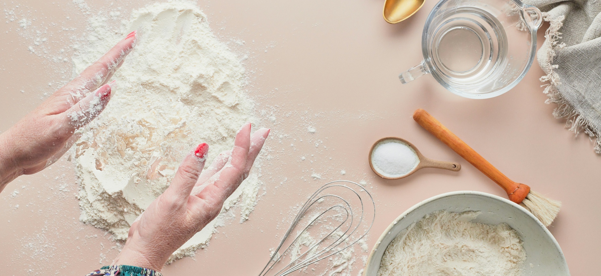 Hände, die mit Mehl arbeiten, liegen auf einer Arbeitsfläche. Um sie herum sind ein Schüssel mit Mehl, ein Messlöffel mit Salz, eine Schüssel mit Wasser, ein Schneebesen und ein Nudelholz zu sehen. Die Hände sind mit Mehl bedeckt und strahlen Vorfreude auf das Backen aus.